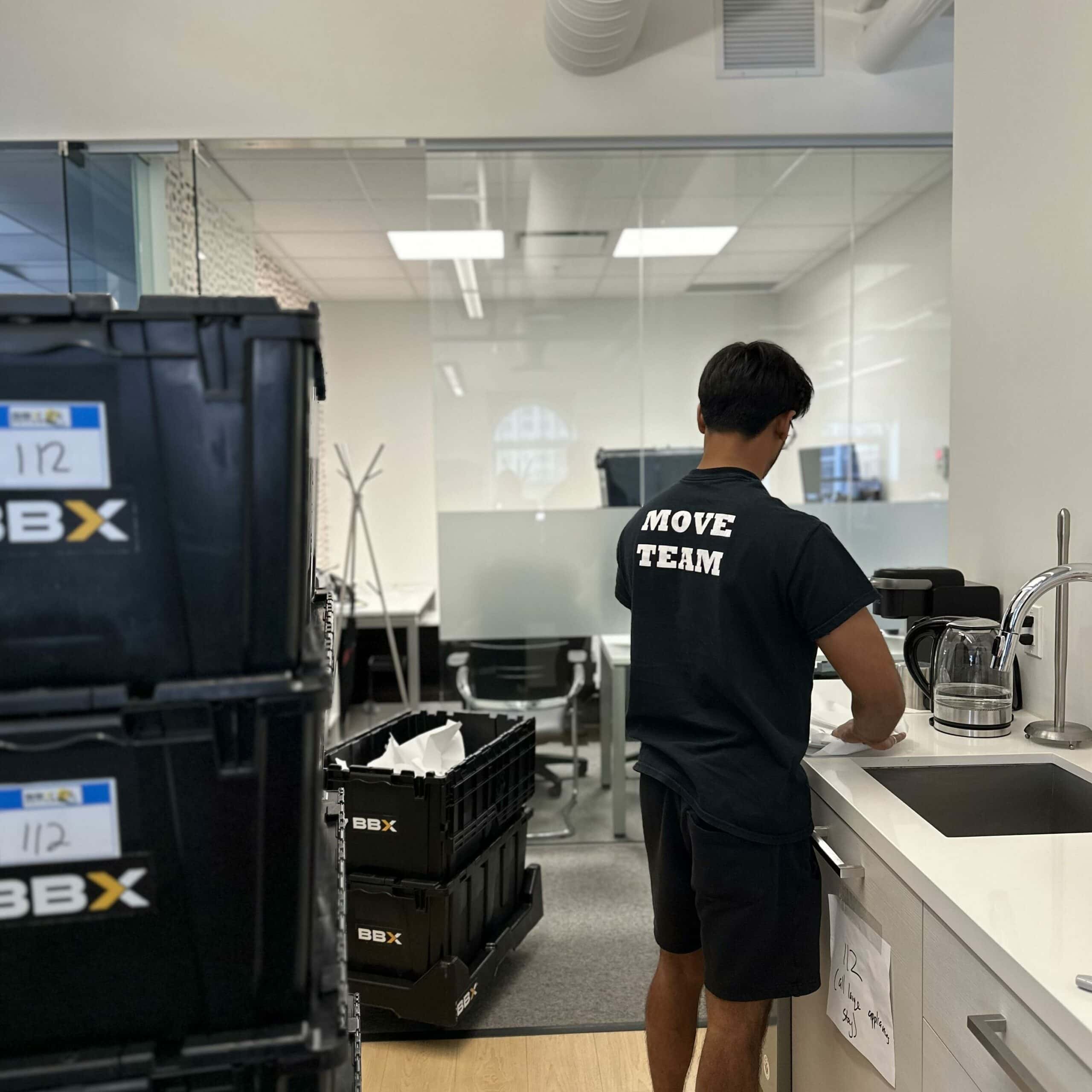 A person in a "Move Team" shirt stands at a kitchen sink in an office, orchestrating the office relocation. Nearby, black crates labeled "BBX" hint at the efficiency of a full-service moving company.