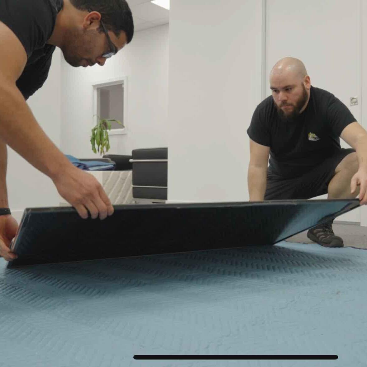 Two men from a full-service moving company are carefully placing a large flat screen TV onto a padded moving blanket in an office relocation setting.