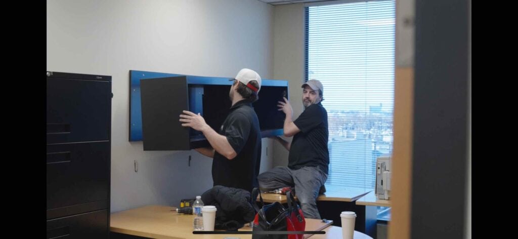 Two men are installing a wall-mounted cabinet in an office space, prepping for office relocation. Tools and beverages sit on the desk nearby, highlighting the efficiency of a full-service moving company.
