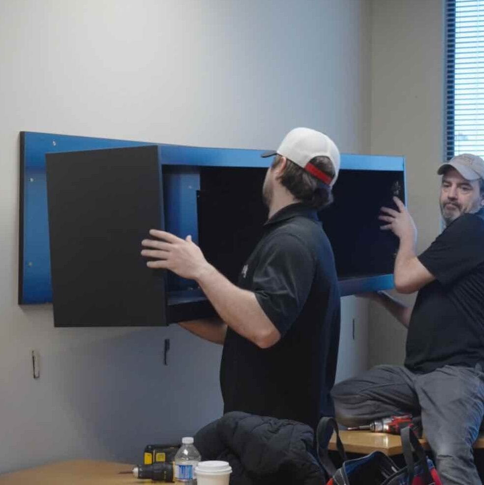 Two men are installing a wall-mounted cabinet in an office space, prepping for office relocation. Tools and beverages sit on the desk nearby, highlighting the efficiency of a full-service moving company.