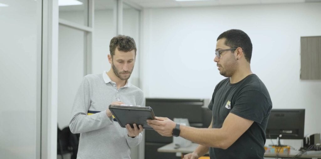 Two men discuss office relocation strategies on a tablet. One wears a grey shirt, the other a black t-shirt, as they review plans provided by a full-service moving company.