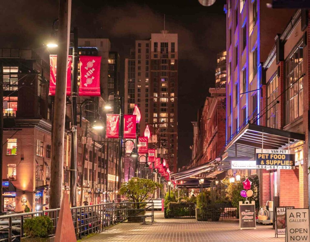 Nighttime street scene in a 2025 Vancouver neighborhood with illuminated buildings, red banners, and shop signs like a pet food store and art gallery. The street is lined with trees and empty benches, creating an inviting urban escape.