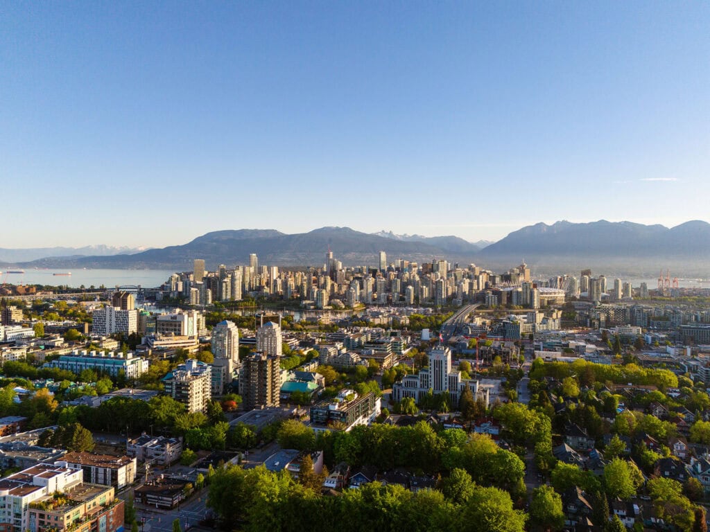 An aerial view of Vancouver's vibrant neighborhoods reveals a cityscape with numerous buildings, lush green trees, and distant mountains under a clear blue sky.