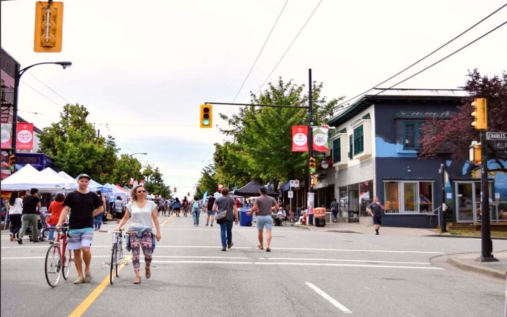 In 2025, Vancouver's neighborhoods come alive as people stroll and bike along streets closed to traffic, lined with vibrant stalls and shops. Banners sway from streetlights while trees provide shade along the bustling sidewalks.