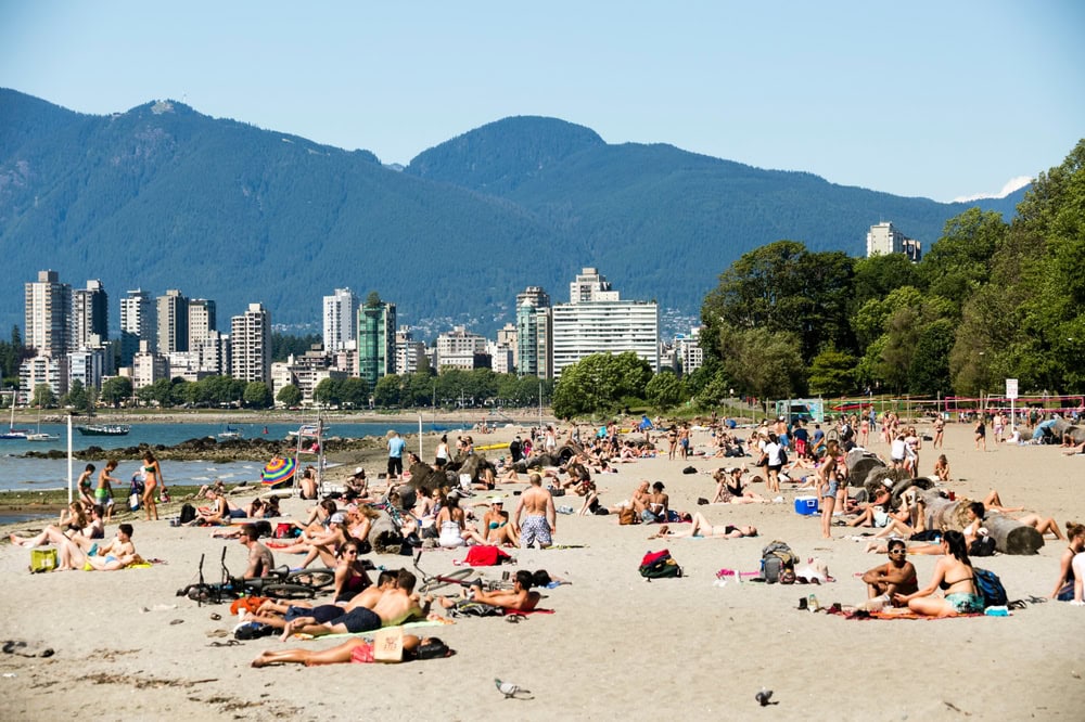 People sunbathing and relaxing on a sandy beach in Vancouver, with its vibrant neighborhoods framing a cityscape against the backdrop of majestic mountains.