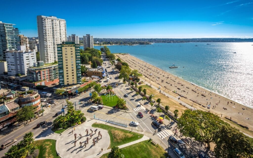 Aerial view of a sandy beach along a busy city street in Vancouver, with tall buildings, lush trees, and vibrant neighborhoods. People are sunbathing while a striking sculpture punctuates the circular plaza area near the road in this scenic 2025 cityscape.