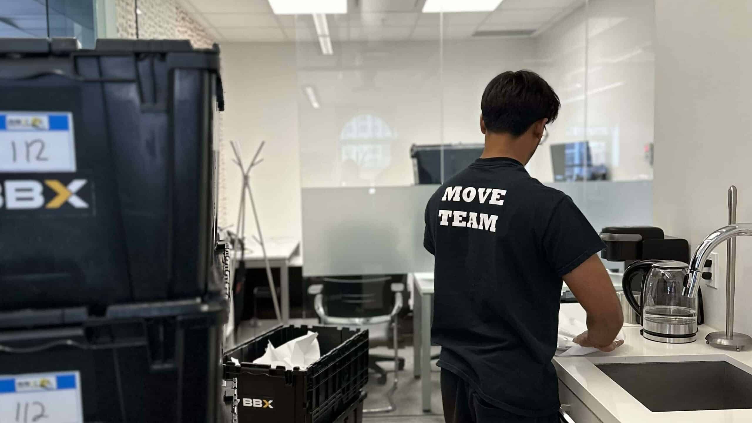A person in a "Move Team" shirt stands at a kitchen sink in an office, orchestrating the office relocation. Nearby, black crates labeled "BBX" hint at the efficiency of a full-service moving company.