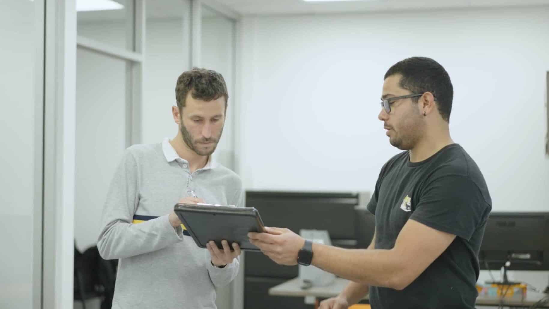 Two men discuss office relocation strategies on a tablet. One wears a grey shirt, the other a black t-shirt, as they review plans provided by a full-service moving company.