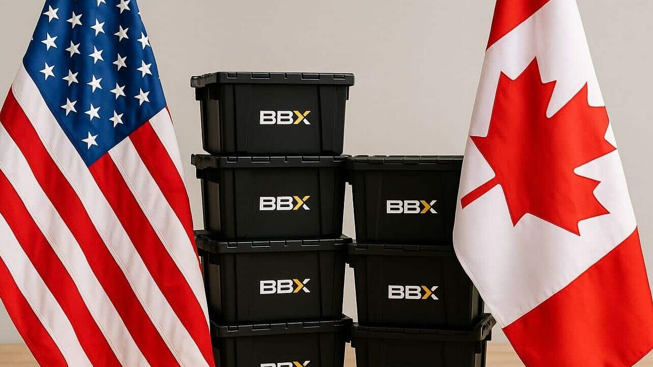 Six black storage boxes labeled "BBX" are stacked on a cart between American and Canadian flags, symbolizing the journey of moving to Vancouver from the US, all set on a wooden surface against a neutral background.