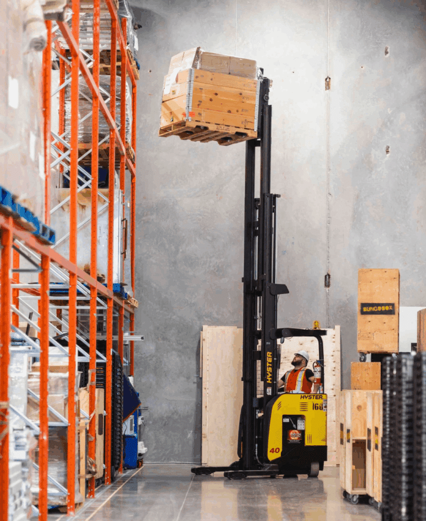 A worker operates a yellow forklift, raising a pallet of wooden crates high onto an industrial warehouse shelf—demonstrating the precision and expertise of Industrial Moving Services.