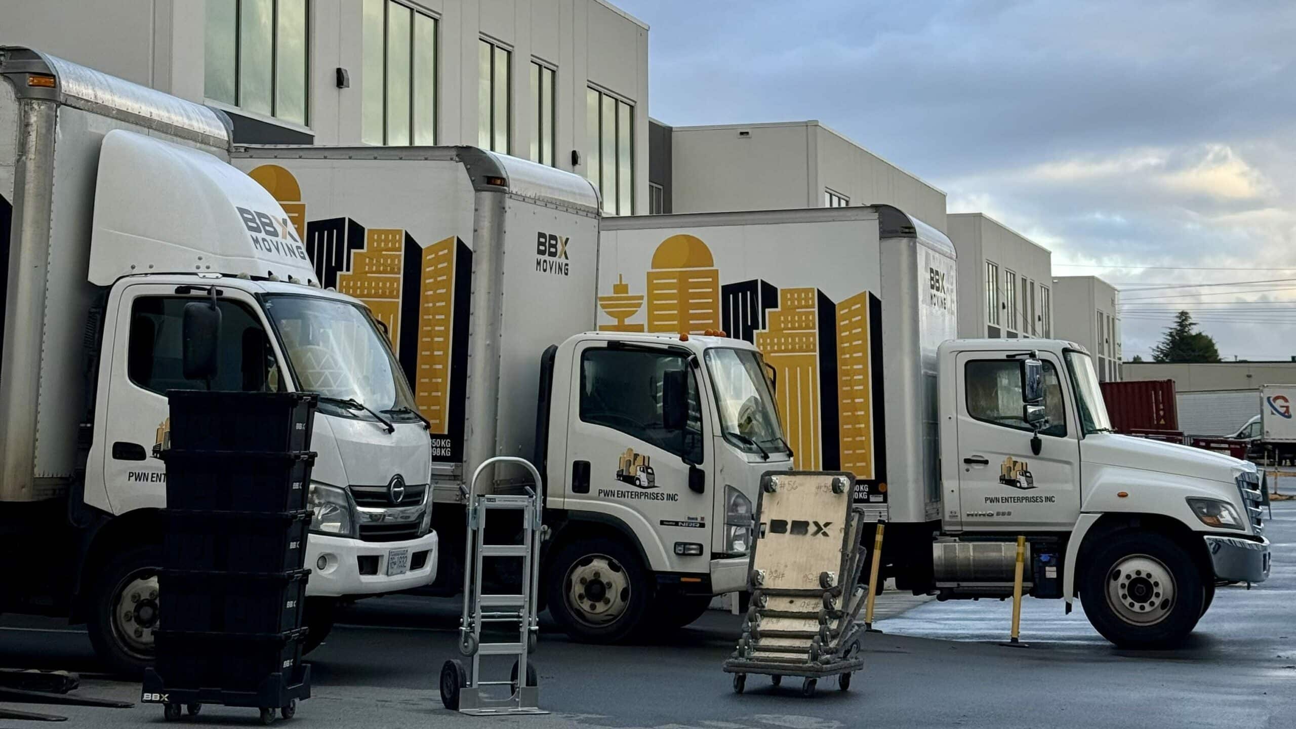 Three delivery trucks are parked outside a modern industrial building, with moving equipment and plastic bins on the wet ground nearby under a cloudy sky—an essential scene for any long-distance move.