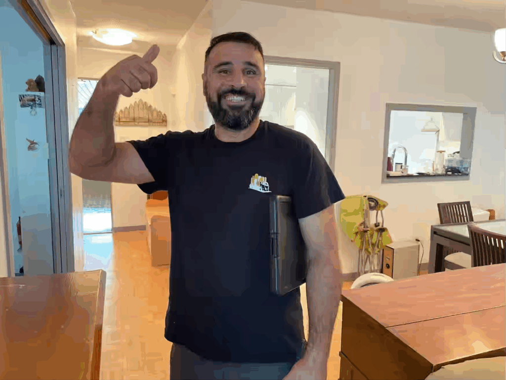 A bearded man stands indoors, smiling and giving a thumbs-up while holding a closed laptop under his arm—ready to share his favorite move day tips in the comfort of his living or dining area.
