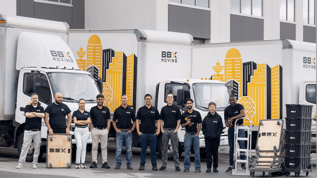 Ten people in matching BBX Moving shirts stand in front of three branded moving trucks and equipment outside a commercial building, ready to deliver a stress-free move.