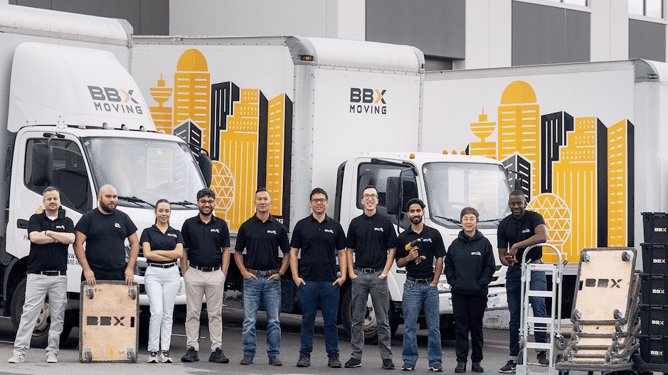 Ten people in matching BBX Moving shirts stand in front of three branded moving trucks and equipment outside a commercial building, ready to deliver a stress-free move.