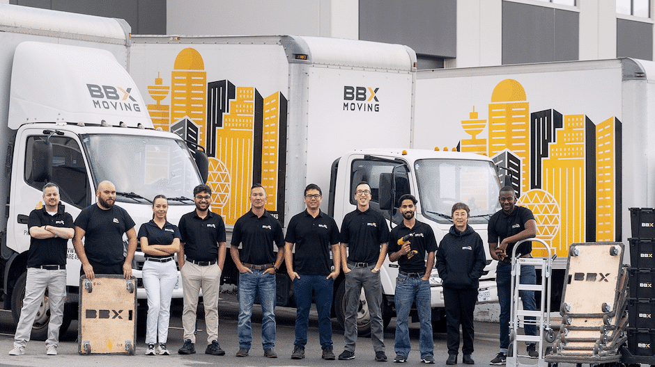 Ten people in matching BBX Moving shirts stand in front of three branded moving trucks and equipment outside a commercial building, ready to deliver a stress-free move.