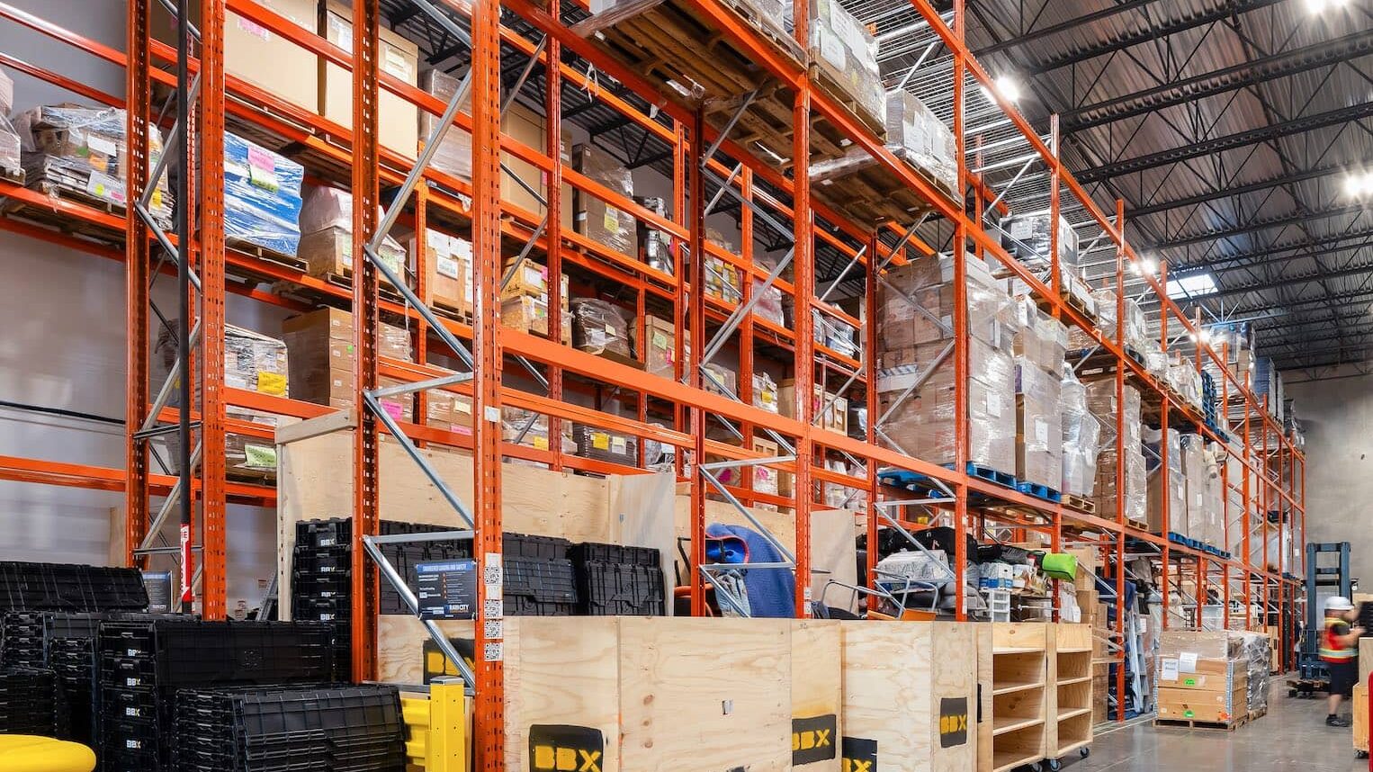 A warehouse. Pallets and crates are neatly organized, perfect for temporary storage needs. The ceiling's metal beams and bright lights illuminate the space efficiently.