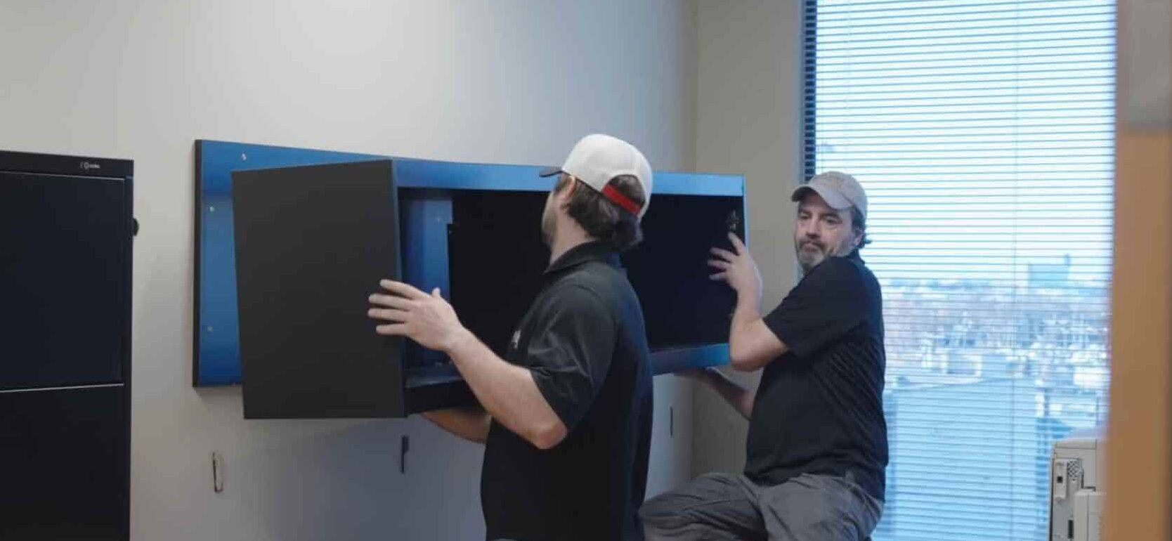 Two men are installing a wall-mounted cabinet in an office space, prepping for office relocation. Tools and beverages sit on the desk nearby, highlighting the efficiency of a full-service moving company.