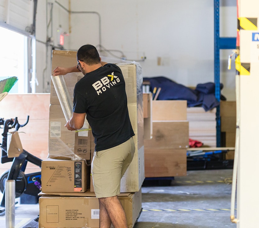 BBX Moving | Commercial Movers – Burnaby - A man wearing a "BBX Moving" shirt wraps plastic around a large box in a Richmond storage or warehouse area, highlighting the expertise of commercial movers surrounded by boxes and equipment. A man wearing a "BBX Moving" shirt wraps plastic around a large box in a Richmond storage or warehouse area, highlighting the expertise of commercial movers surrounded by boxes and equipment.