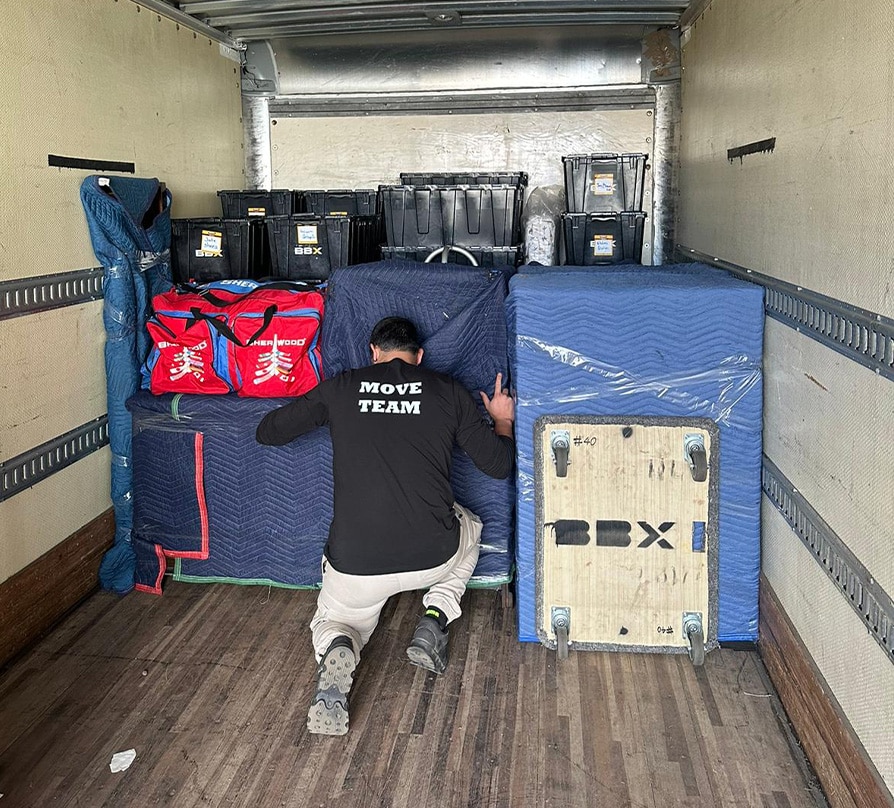 BBX Moving | Commercial Movers – Burnaby - A person wearing a "MOVE TEAM" shirt kneels in a moving truck, organizing wrapped furniture, boxes, red bags, and plastic bins—hard at work with experienced Richmond Movers. A person wearing a "MOVE TEAM" shirt kneels in a moving truck, organizing wrapped furniture, boxes, red bags, and plastic bins—hard at work with experienced Richmond Movers.