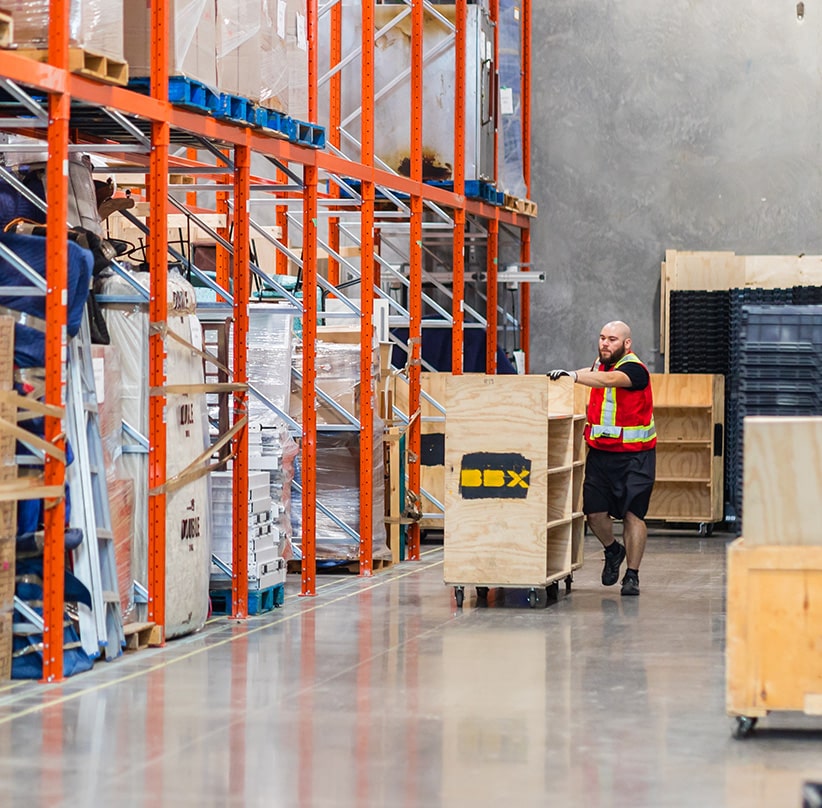 BBX Moving | Commercial Movers – Burnaby - A worker in a safety vest moves a large wooden crate on wheels through a Richmond warehouse with tall orange storage racks, showcasing the efficiency of commercial movers. A worker in a safety vest moves a large wooden crate on wheels through a Richmond warehouse with tall orange storage racks, showcasing the efficiency of commercial movers.