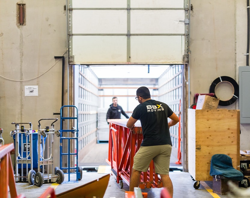 BBX Moving | Commercial Movers – Burnaby - Two men, expert Commercial Movers, load a large red metal structure onto a moving truck inside a Richmond warehouse. One wears a "BBX Moving" shirt and pushes the item toward the truck's open cargo area. Two men, expert Commercial Movers, load a large red metal structure onto a moving truck inside a Richmond warehouse. One wears a "BBX Moving" shirt and pushes the item toward the truck's open cargo area.