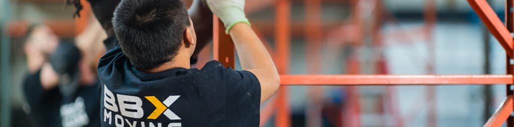 BBX Moving | Warehouse Movers – West Vancouver - A person in a "BBX Moving" shirt lifts or assembles an orange metal structure indoors, likely as part of Warehouse Movers work in Richmond. Other people are visible in the blurred background. A person in a "BBX Moving" shirt lifts or assembles an orange metal structure indoors, likely as part of Warehouse Movers work in Richmond. Other people are visible in the blurred background.