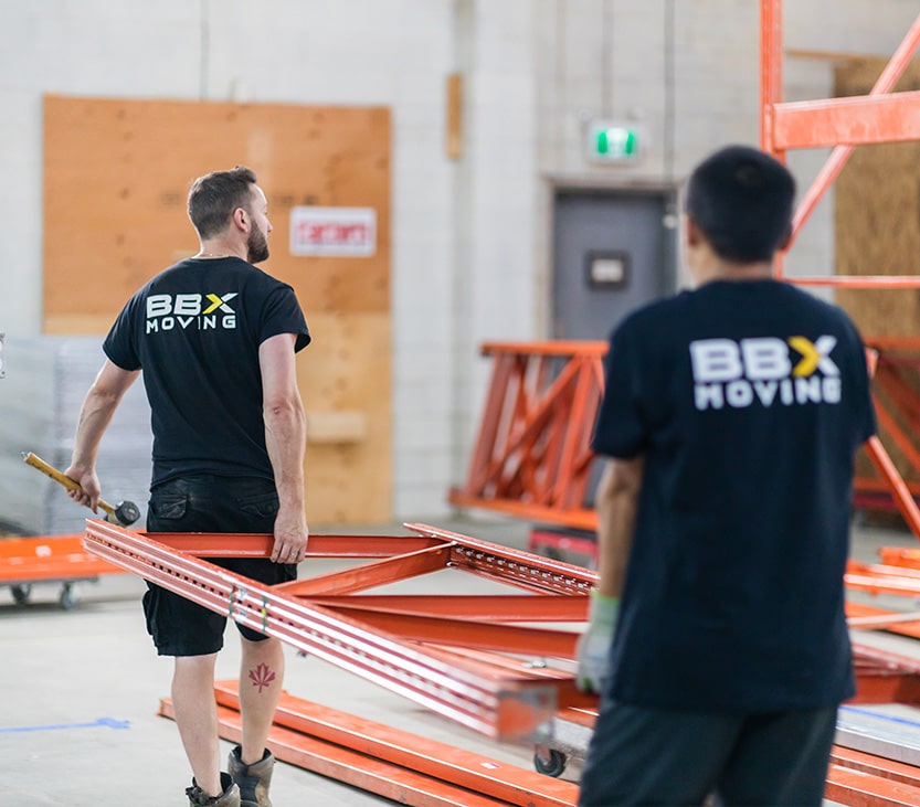 BBX Moving | Warehouse Movers – West Vancouver - Two workers wearing "BBX Moving" shirts, experienced warehouse movers, carry a large orange metal frame inside a Richmond warehouse with construction materials in the background. Two workers wearing "BBX Moving" shirts, experienced warehouse movers, carry a large orange metal frame inside a Richmond warehouse with construction materials in the background.