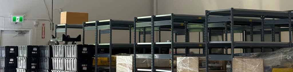 BBX Moving | Commercial Movers – Burnaby - Rows of empty metal shelving units and stacked black storage bins in a Richmond warehouse with cardboard boxes, a visible emergency exit sign, and preparation by commercial movers. Rows of empty metal shelving units and stacked black storage bins in a Richmond warehouse with cardboard boxes, a visible emergency exit sign, and preparation by commercial movers.