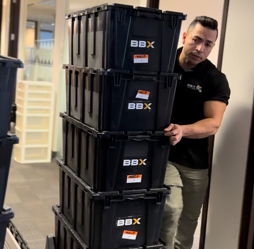 BBX Moving | Office Movers – Burnaby - A man in a black shirt moves a tall stack of four black BBX labeled storage bins through an office hallway with Richmond Office Movers. A man in a black shirt moves a tall stack of four black BBX labeled storage bins through an office hallway with Richmond Office Movers.