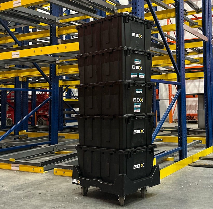 BBX Moving | Warehouse Movers – West Vancouver - Four stacked black BBX storage containers on a wheeled cart sit in front of empty yellow and blue shelves, ready for warehouse relocation with experienced Richmond movers. Four stacked black BBX storage containers on a wheeled cart sit in front of empty yellow and blue shelves, ready for warehouse relocation with experienced Richmond movers.