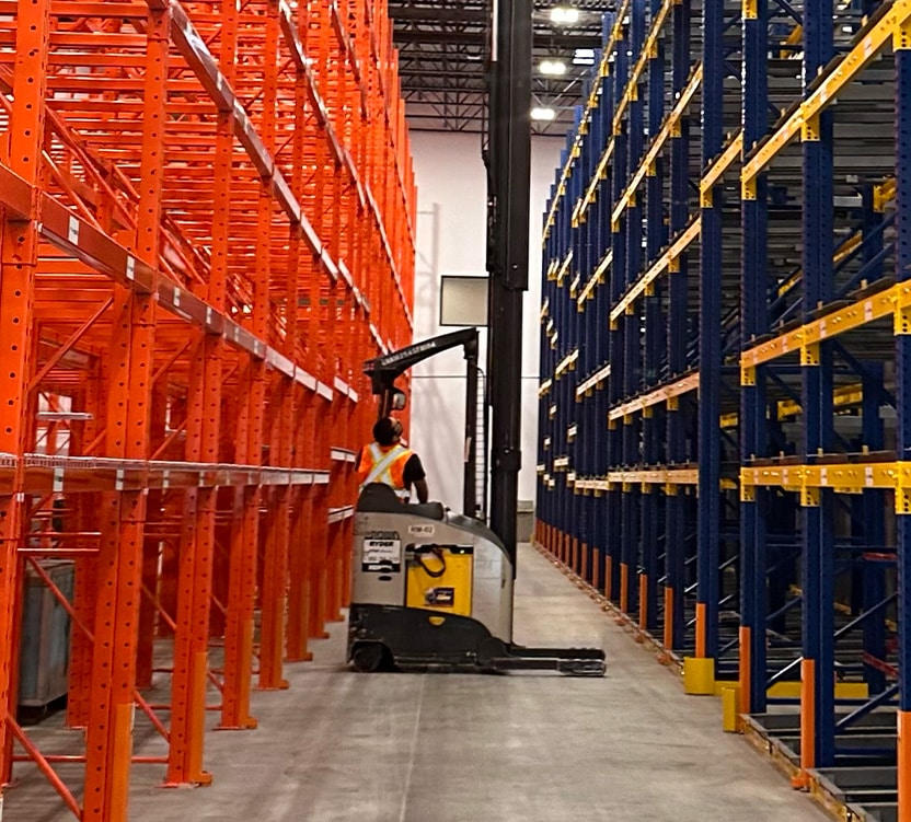 BBX Moving | Warehouse Movers – West Vancouver - A Warehouse Movers employee operates a forklift between empty orange and blue storage racks inside a large industrial facility in Richmond. A Warehouse Movers employee operates a forklift between empty orange and blue storage racks inside a large industrial facility in Richmond.