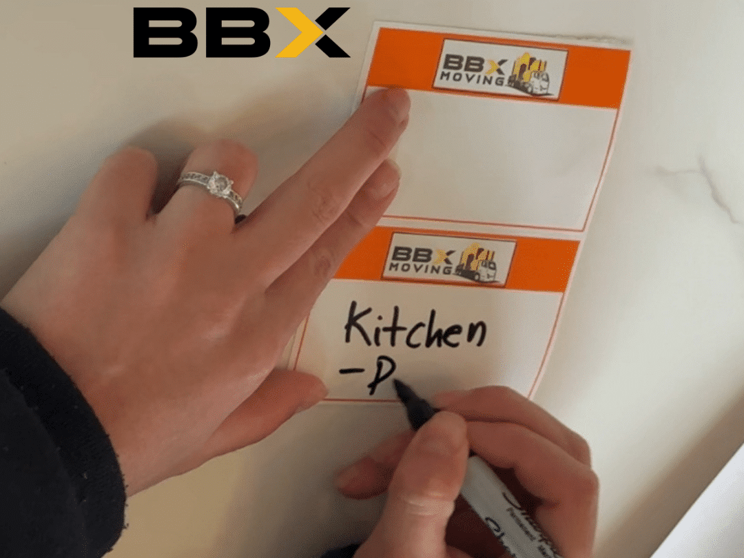 BBX Moving | How to Organize a Residential Move | BBX Moving - A person writes "Kitchen" on a BBX Moving label with a black marker, taking the first step to organize a move seamlessly. Text above reads "Care that shows. BBX. A person writes "Kitchen" on a BBX Moving label with a black marker, taking the first step to organize a move seamlessly. Text above reads "Care that shows. BBX.