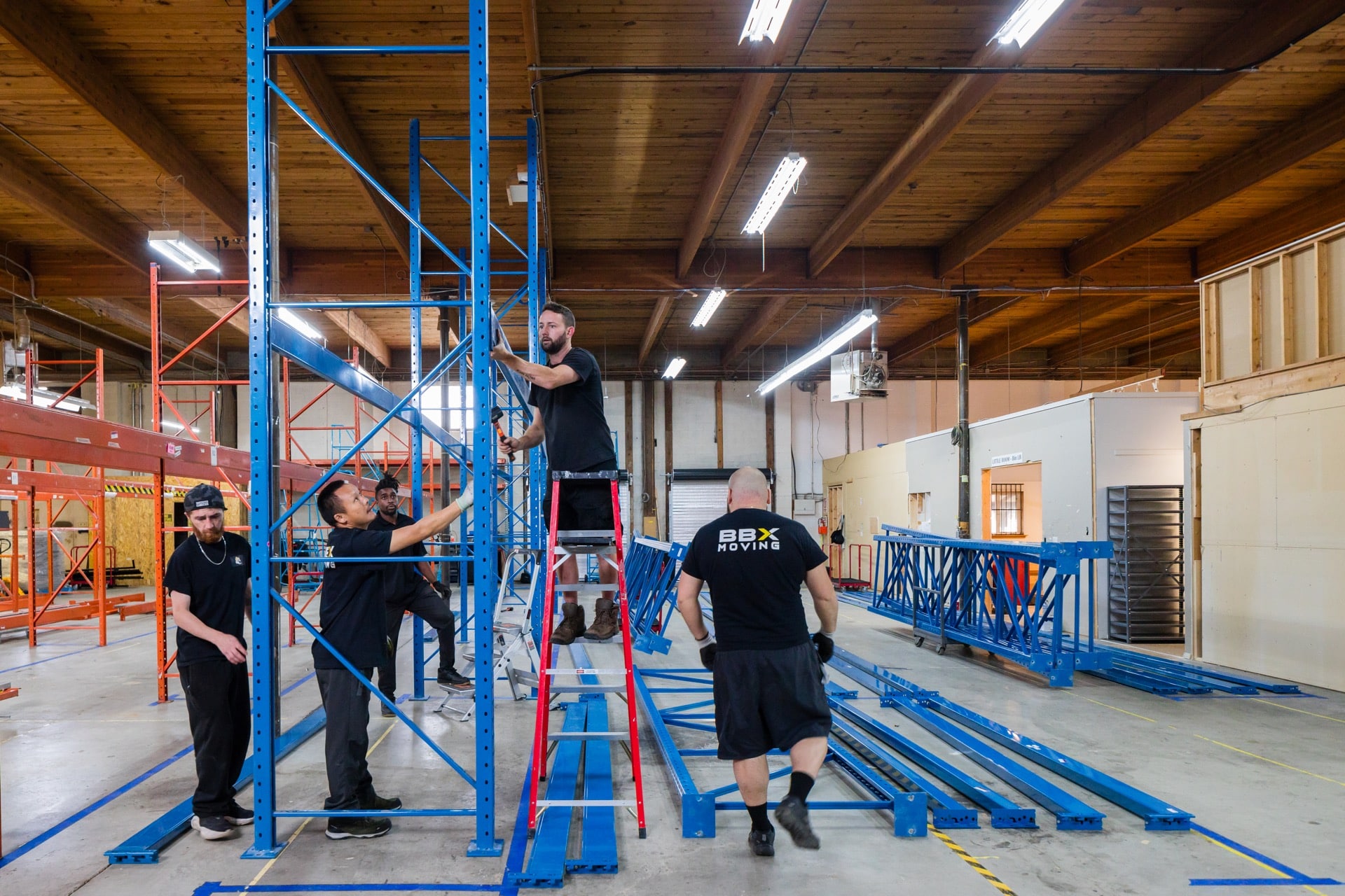 BBX Moving | Locations - Five people assemble blue metal shelving units in a warehouse, preparing the space for incoming storage needs commonly required by moving companies in the Lower Mainland. One person is on a ladder, while others hold and position the pieces. The space has exposed wooden beams and fluorescent lighting. Five people assemble blue metal shelving units in a warehouse, preparing the space for incoming storage needs commonly required by moving companies in the Lower Mainland. One person is on a ladder, while others hold and position the pieces. The space has exposed wooden beams and fluorescent lighting.