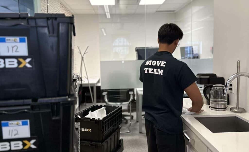BBX Moving | Do I Need To Empty Drawers For Movers On Moving Day? - A person in a "Move Team" shirt stands at a kitchen sink in an office, orchestrating the office relocation. Nearby, black crates labeled "BBX" hint at the efficiency of a full-service moving company. A person in a "Move Team" shirt stands at a kitchen sink in an office, orchestrating the office relocation. Nearby, black crates labeled "BBX" hint at the efficiency of a full-service moving company.